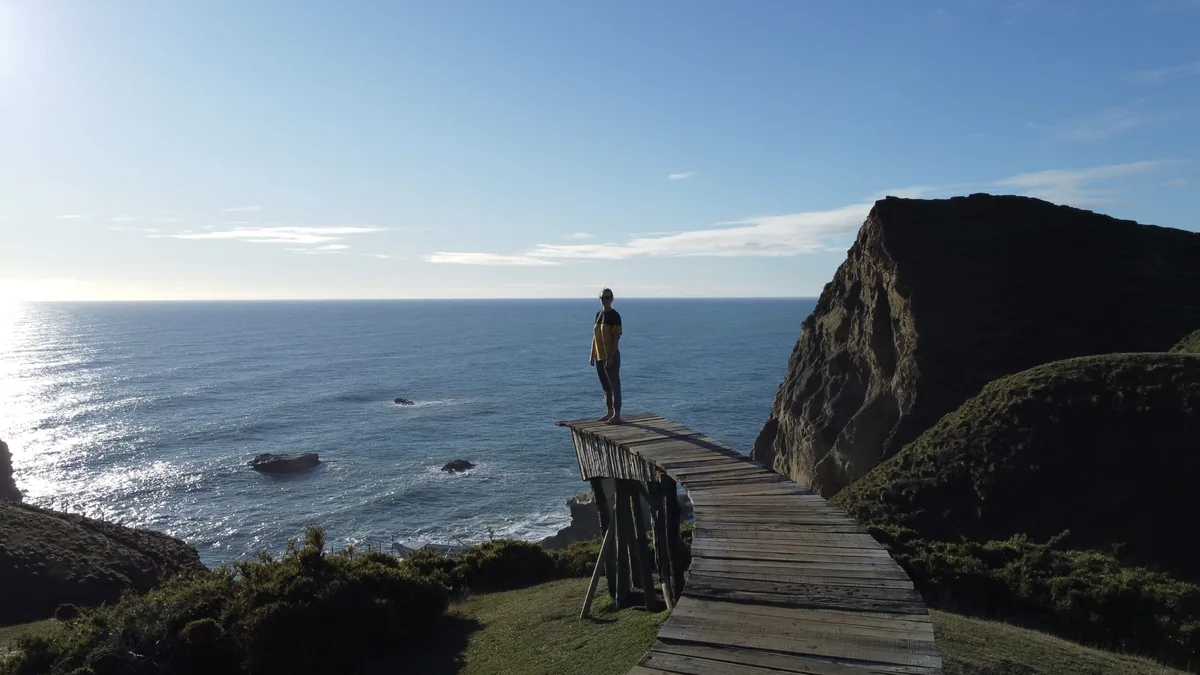 Viajero en mirador de madera sobre acantilado costero, contemplando el vasto océano bajo un sol brillante y cielo azul