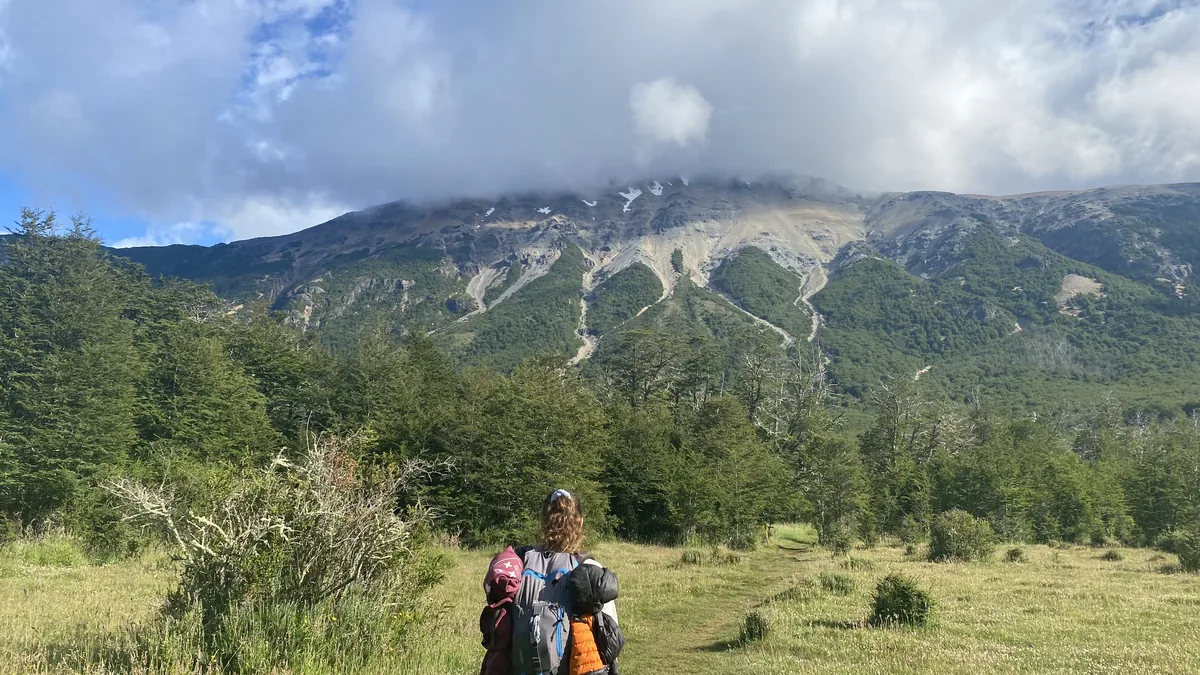 Senderista con mochila grande en un sendero, avanzando hacia montañas boscosas con cumbres rocosas y nevadas parciales