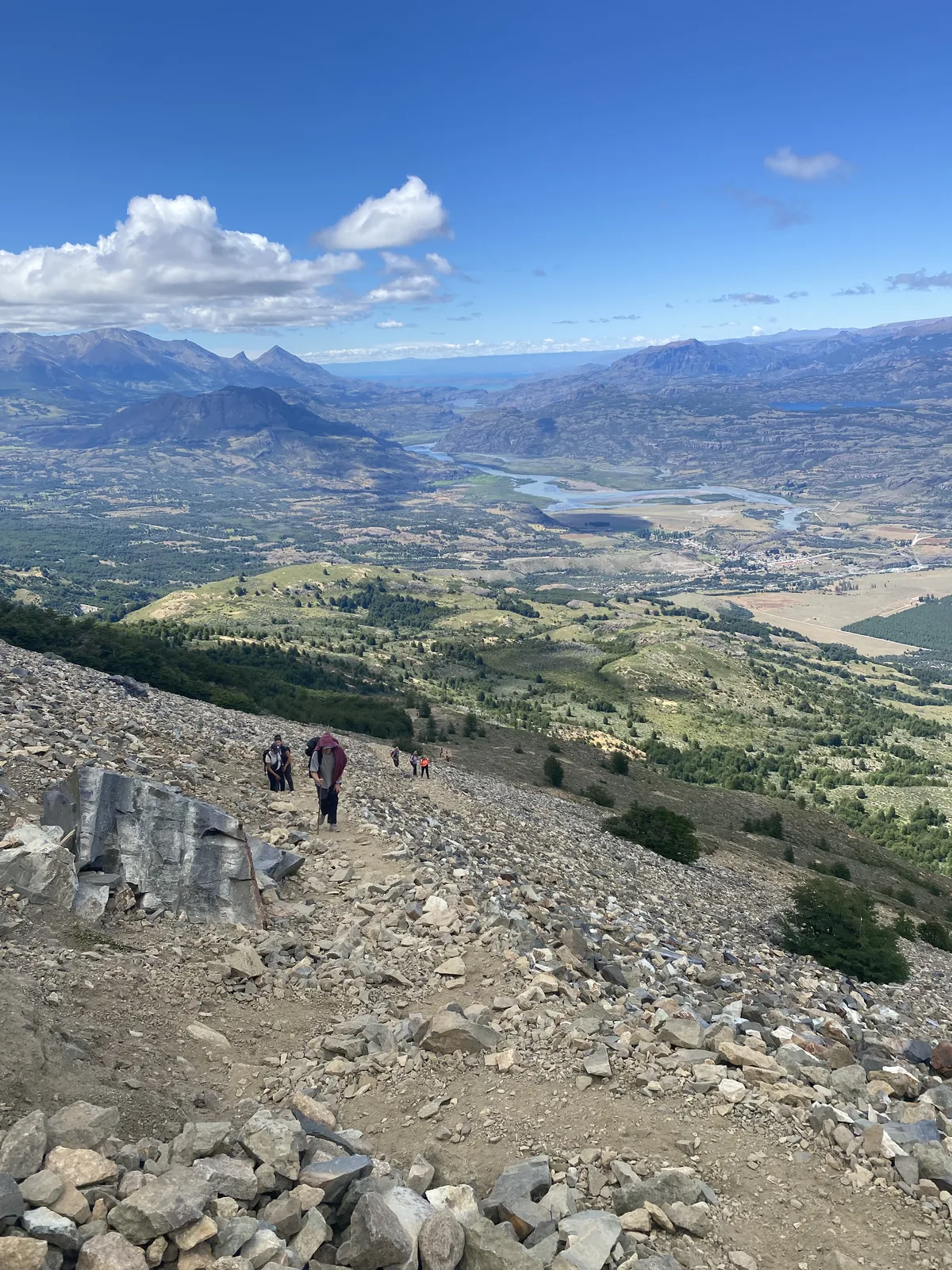 Grupo de senderistas asciende por sendero rocoso, con vistas panorámicas a un vasto valle fluvial y montañas en Patagonia