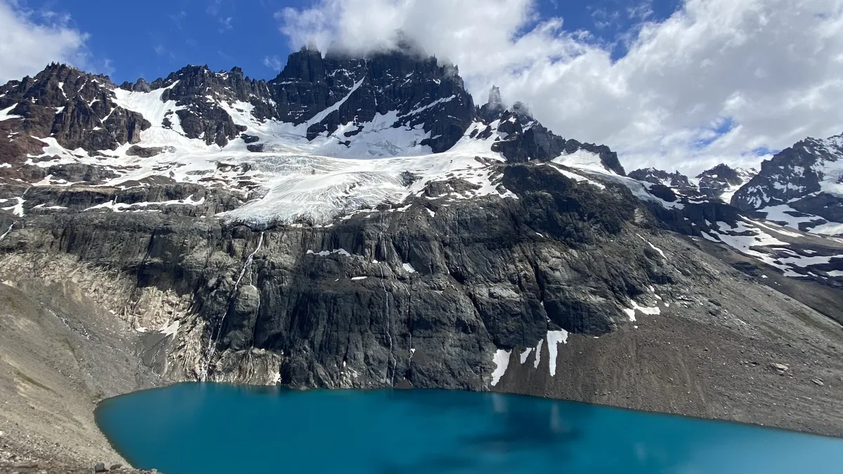 Impresionante lago glacial turquesa al pie de montañas andinas rocosas con glaciares y cumbres nevadas bajo nubes blancas