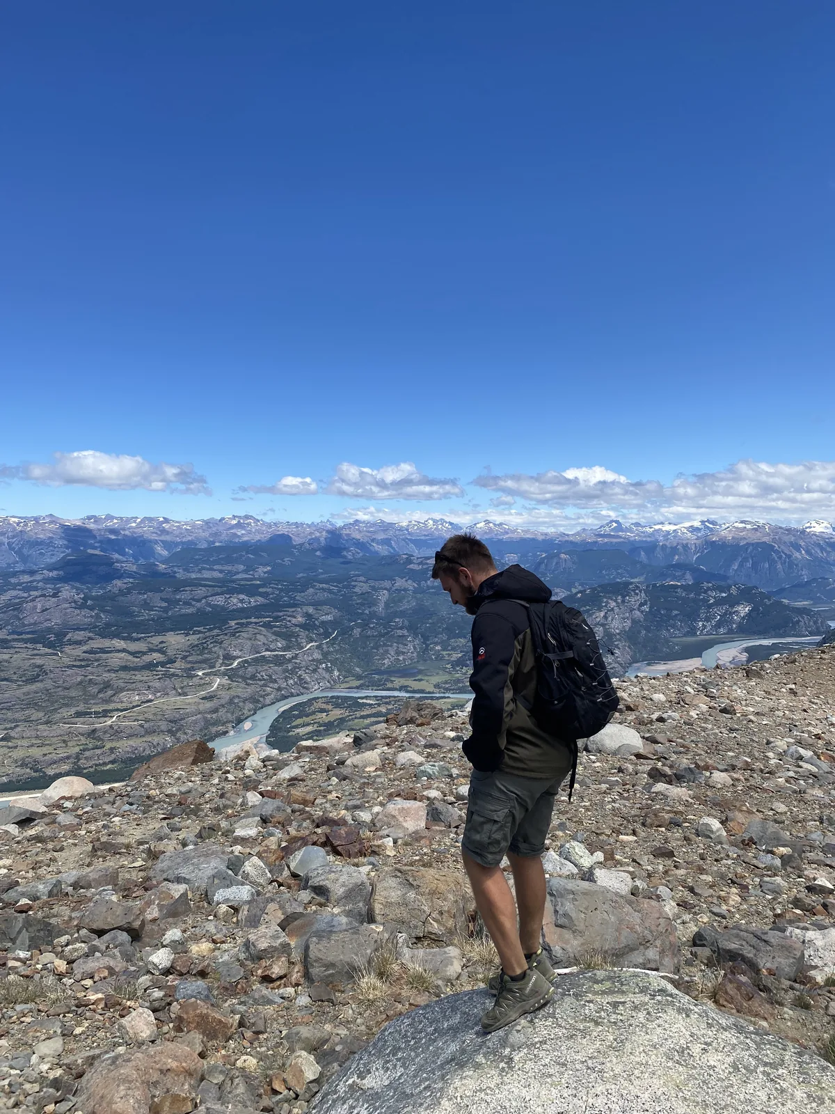 Hombre con mochila y botas de trekking en cima rocosa, explorando paisaje montañoso con río serpenteante y picos nevados