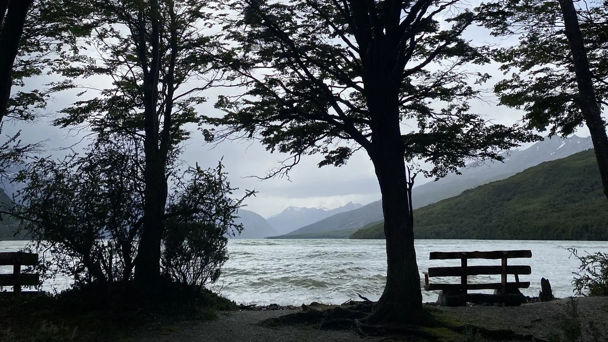 Dos bancos de madera a orillas de un lago agitado, rodeado de montañas nevadas y bosque oscuro bajo un cielo nublado