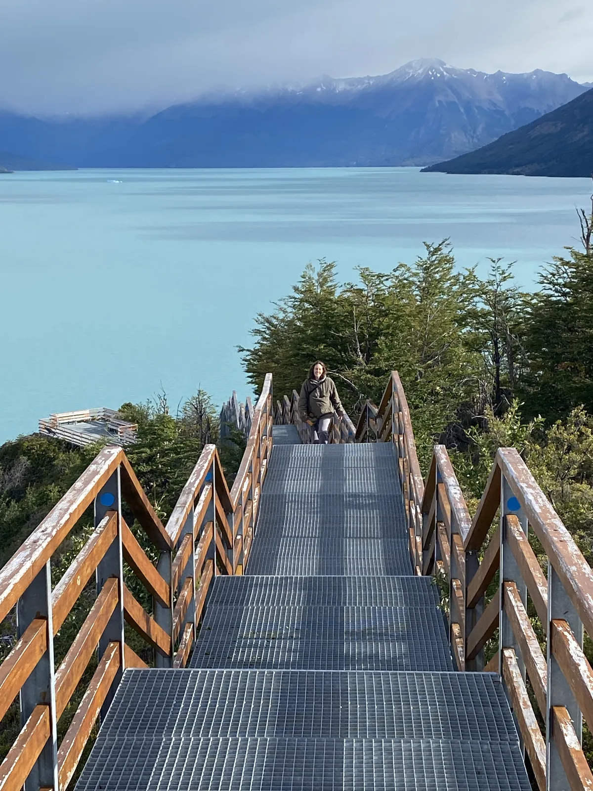 Mujer subiendo una larga escalera metálica con barandales de madera frente a un lago glaciar turquesa y montañas en Patagonia