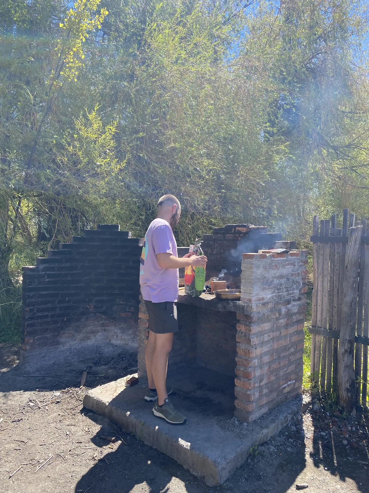 Viajero preparando una barbacoa de ladrillo en un área de camping, rodeado de vegetación verde, disfrutando del sol