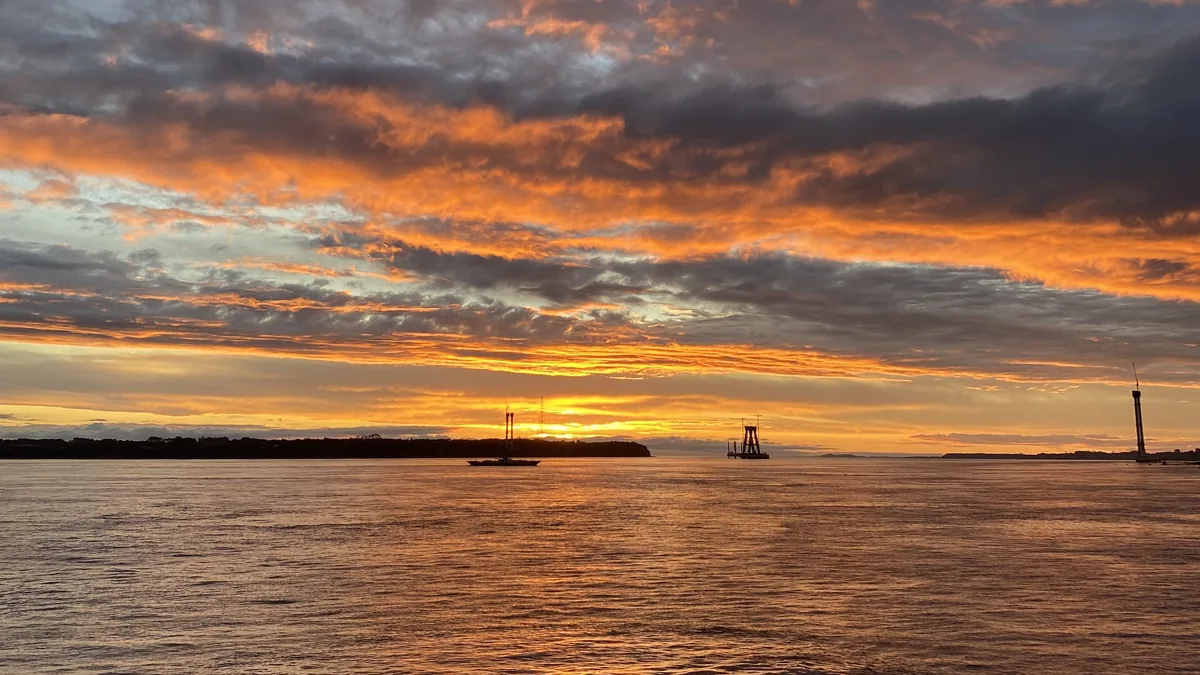 Atardecer vibrante con nubes naranjas sobre un río, un velero y estructuras de puente en construcción en el horizonte