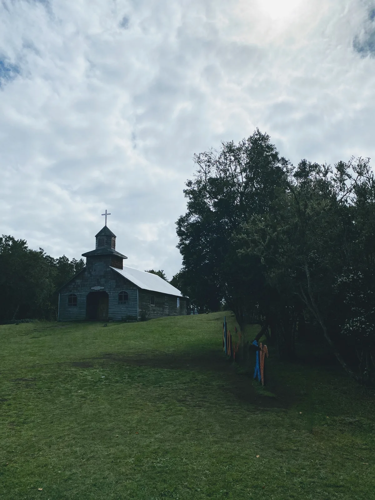 Iglesia rural de madera con cruz en el campanario sobre un campo verde, rodeada de árboles, parada en ruta overland