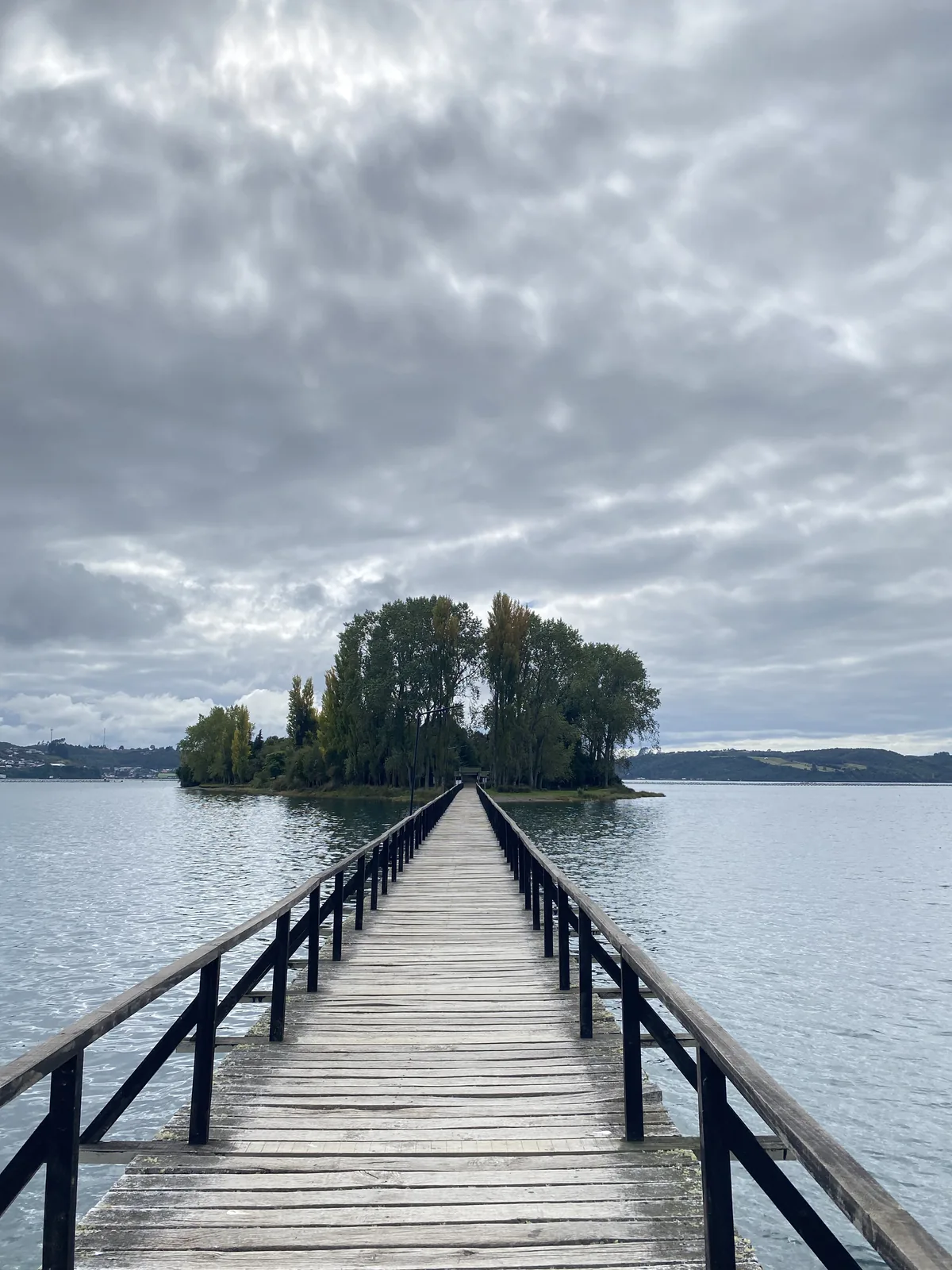 Largo muelle de madera sobre un lago, dirigiendo a una pequeña isla arbolada bajo un cielo nublado
