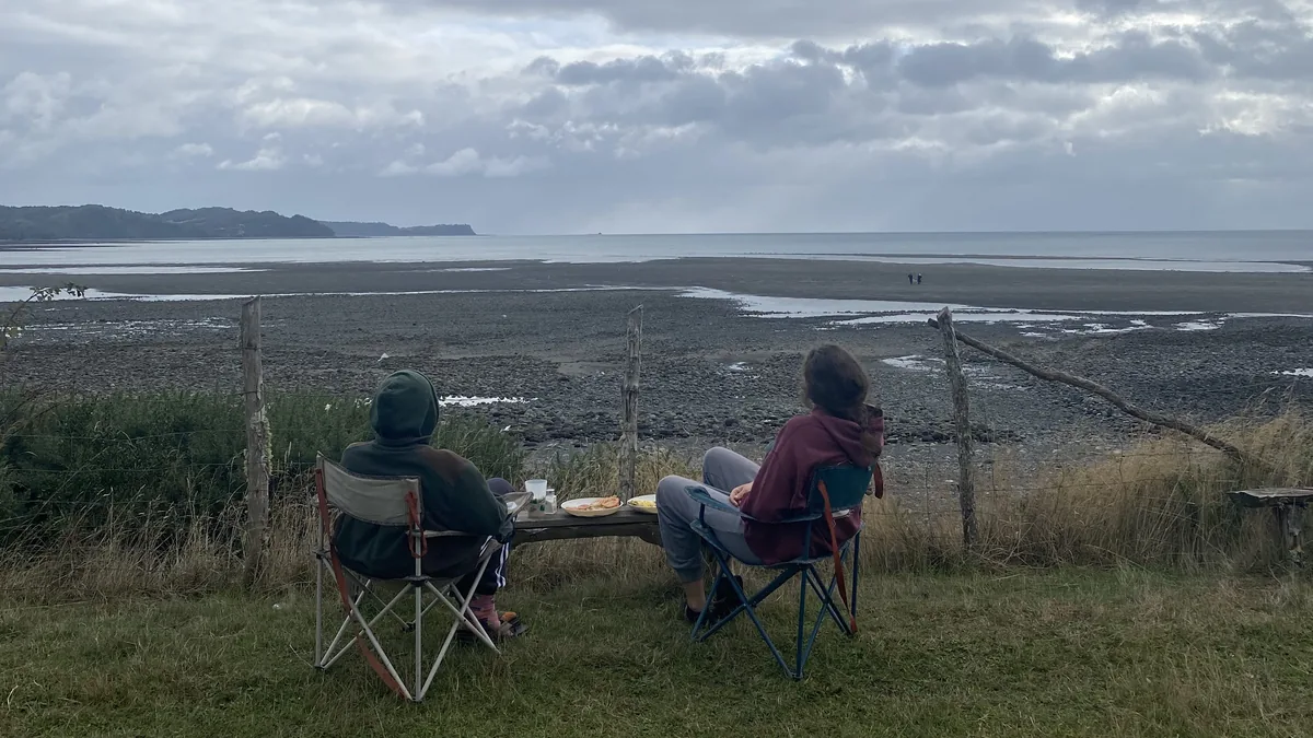 Viajeros overland en sillas de camping disfrutando un picnic junto a una bahía con marea baja y cielo nublado