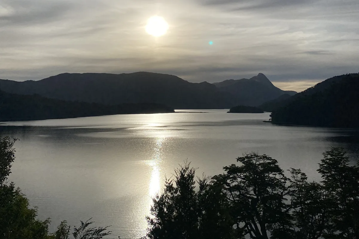 Lago de montaña al atardecer, con el sol reflejado en el agua y siluetas de montañas y árboles en primer plano
