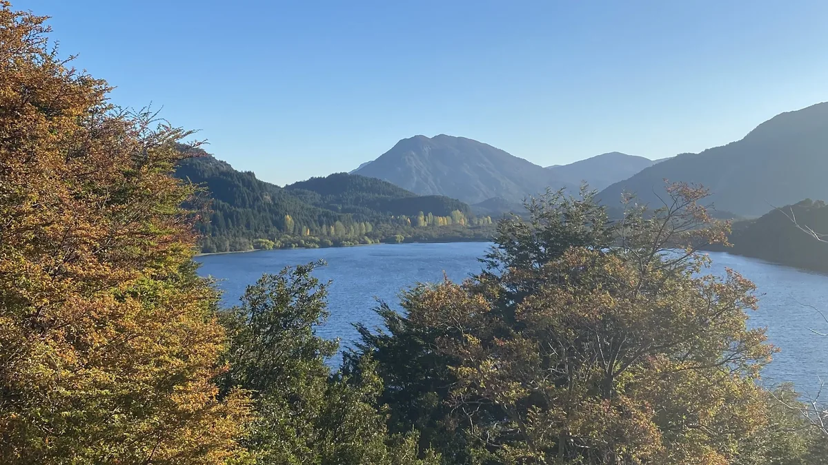 Lago azul profundo entre montañas boscosas, con árboles en primer plano luciendo colores otoñales bajo un cielo despejado