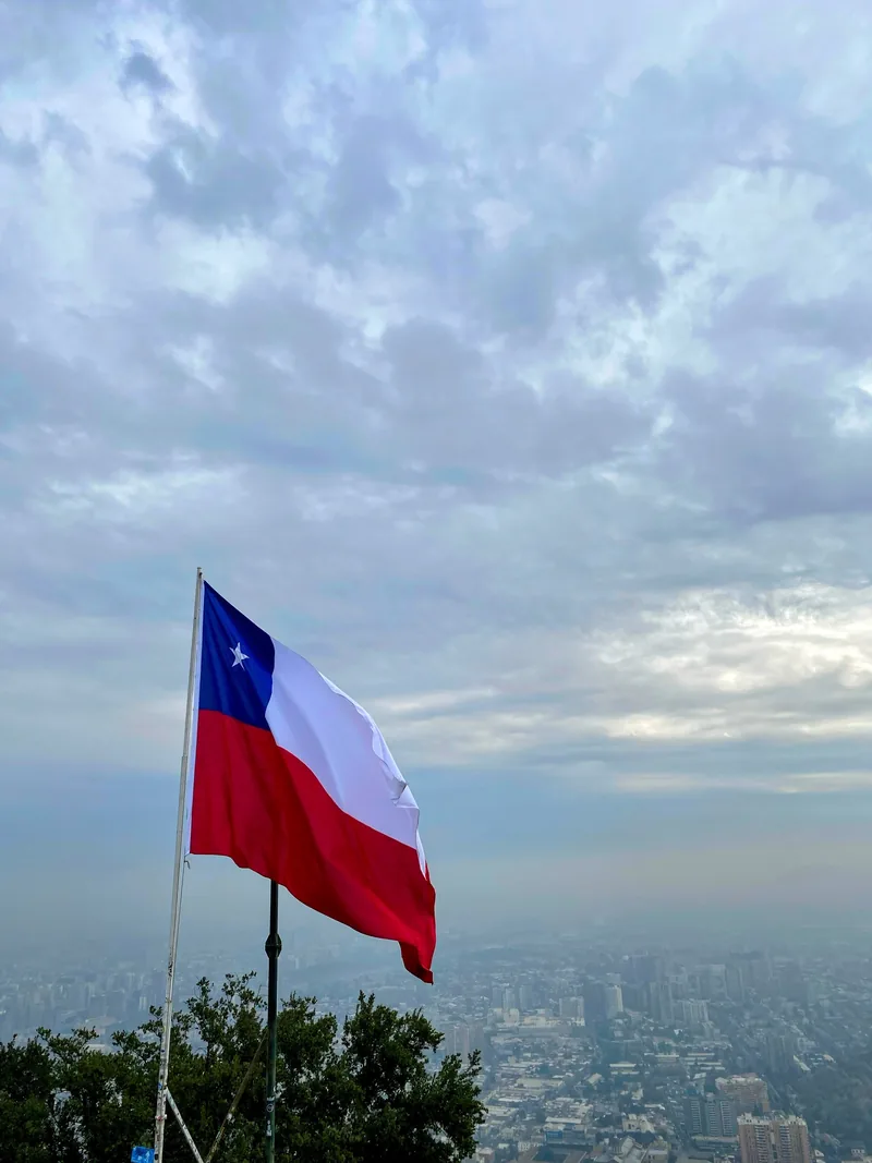 Bandera de Chile flameando sobre Santiago de Chile con la ciudad cubierta de neblina y cielo gris, ruta overland
