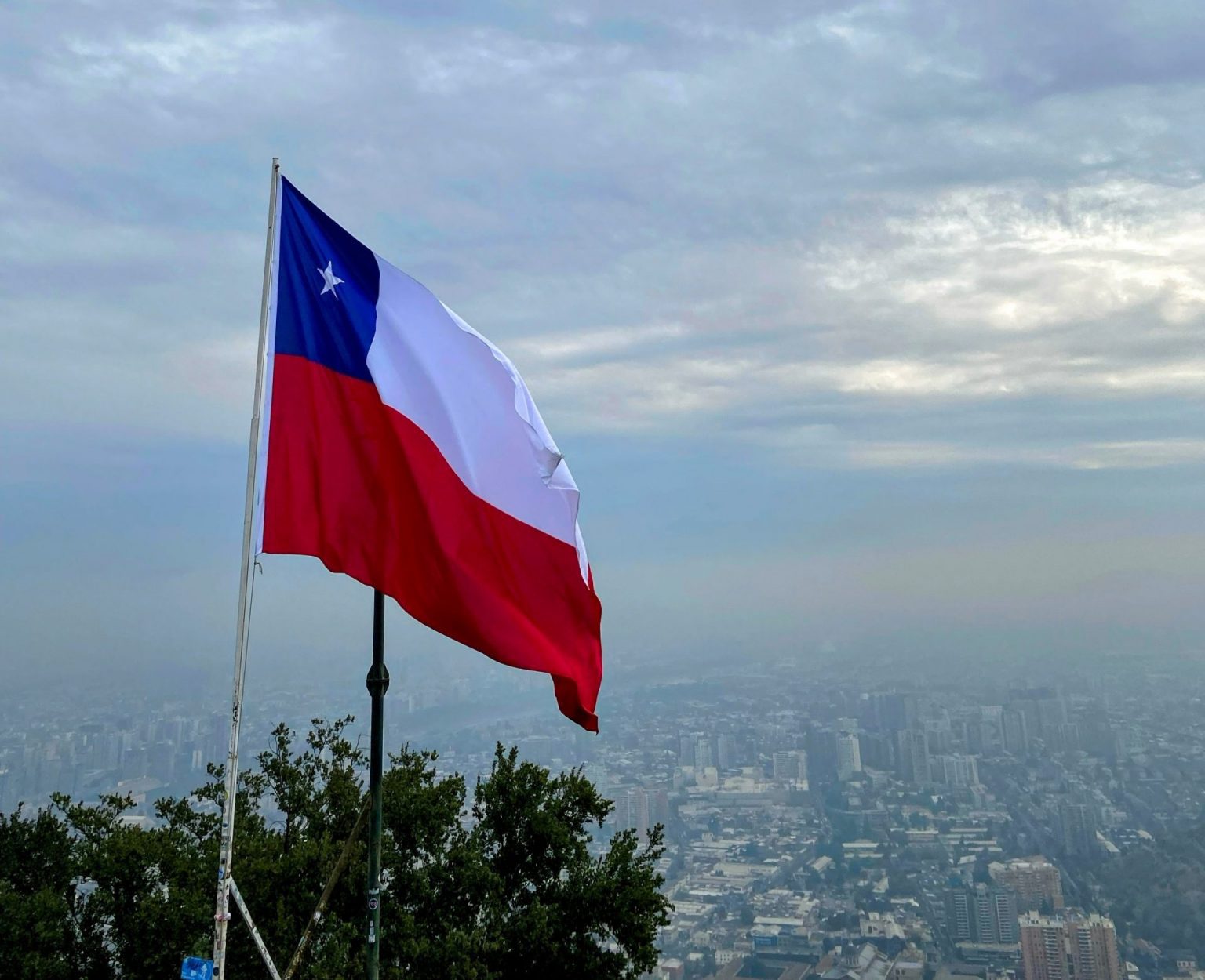 Bandera de Chile flameando sobre Santiago de Chile con la ciudad cubierta de neblina y cielo gris, ruta overland