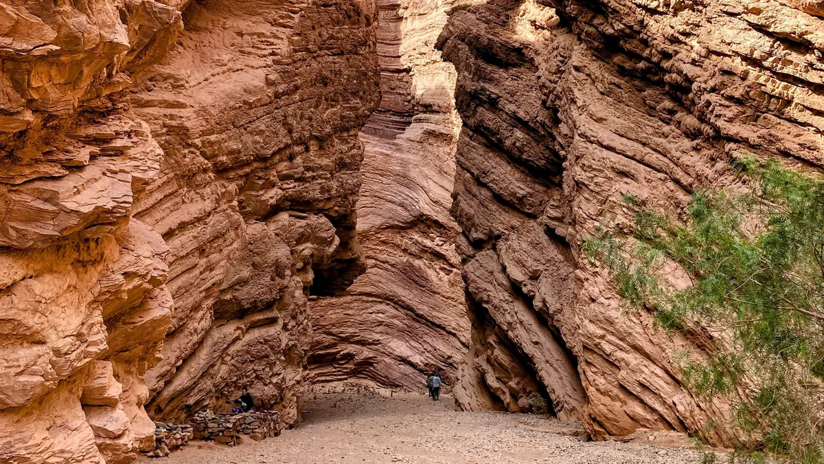 Dos personas exploran un cañón angosto de paredes rocosas estratificadas rojizas, típico de rutas overland andinas