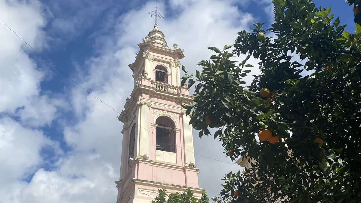 Detallado campanario rosado y blanco de iglesia bajo cielo azul con nubes, junto a un árbol cargado de naranjas vibrantes