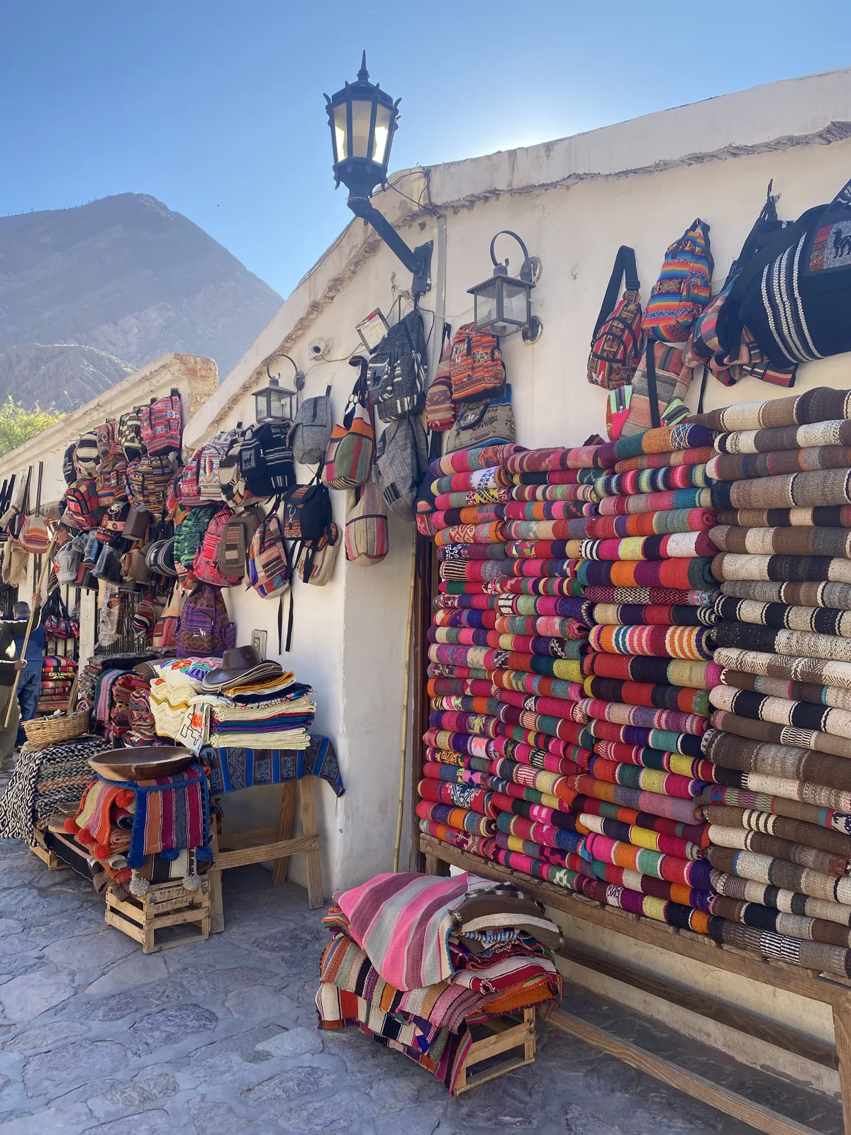 Coloridas mantas y bolsos artesanales en un mercado andino, con montañas bajo el cielo azul de Jujuy