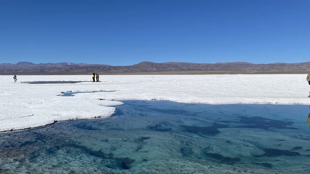 Salar blanco con charcas de agua cristalina bajo un cielo azul intenso y montañas a lo lejos. Viajeros disfrutan del paisaje