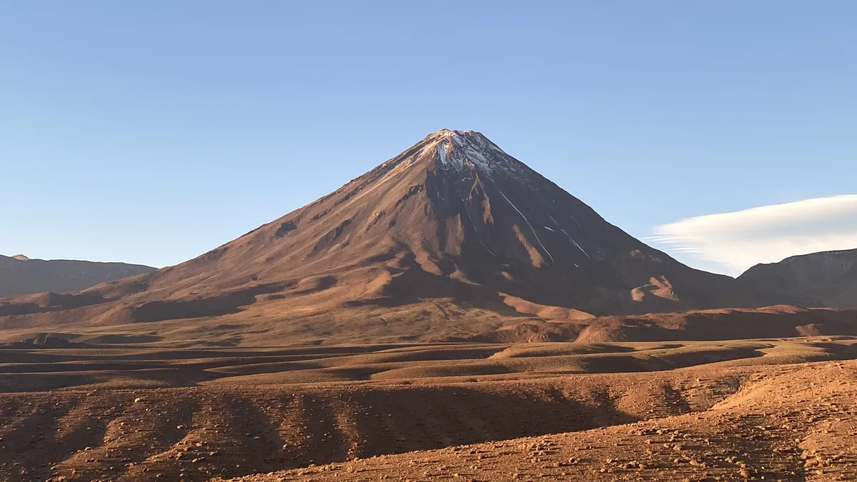 Volcán andino nevado emergiendo de un paisaje desértico rojizo y árido bajo el cielo azul del altiplano