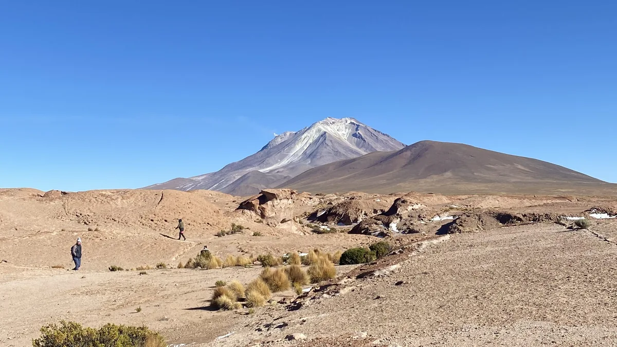 Overlanders caminan por el desierto andino, con formaciones rocosas y un majestuoso volcán de cumbre nevada