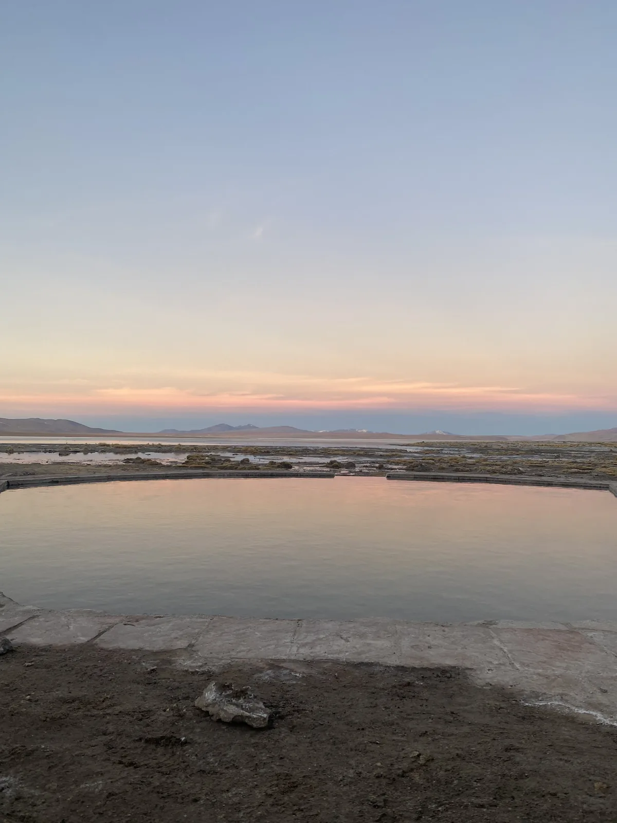 Cielos pastel de atardecer sobre termas naturales en un vasto paisaje altiplánico con montañas al fondo