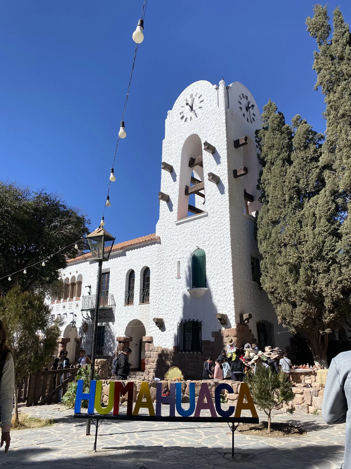 Campanario blanco con relojes y letrero HUMAHUACA en la Quebrada de Jujuy, rodeado de gente y árboles bajo el sol