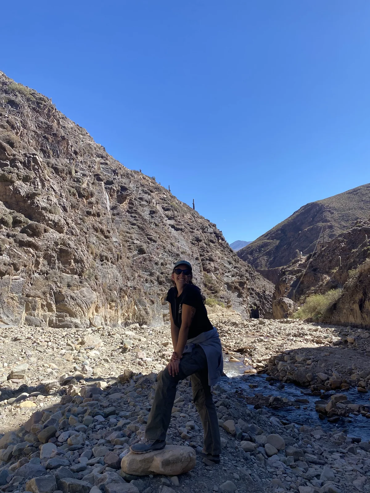 Viajera feliz posando en un cañón rocoso con un riachuelo, montañas áridas y cactáceas bajo el sol