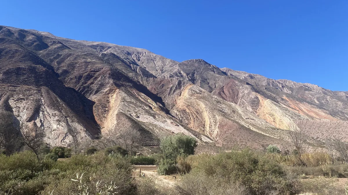 Imponente paisaje andino de montañas con capas de roca multicolores y vegetación arbustiva bajo un cielo azul vibrante