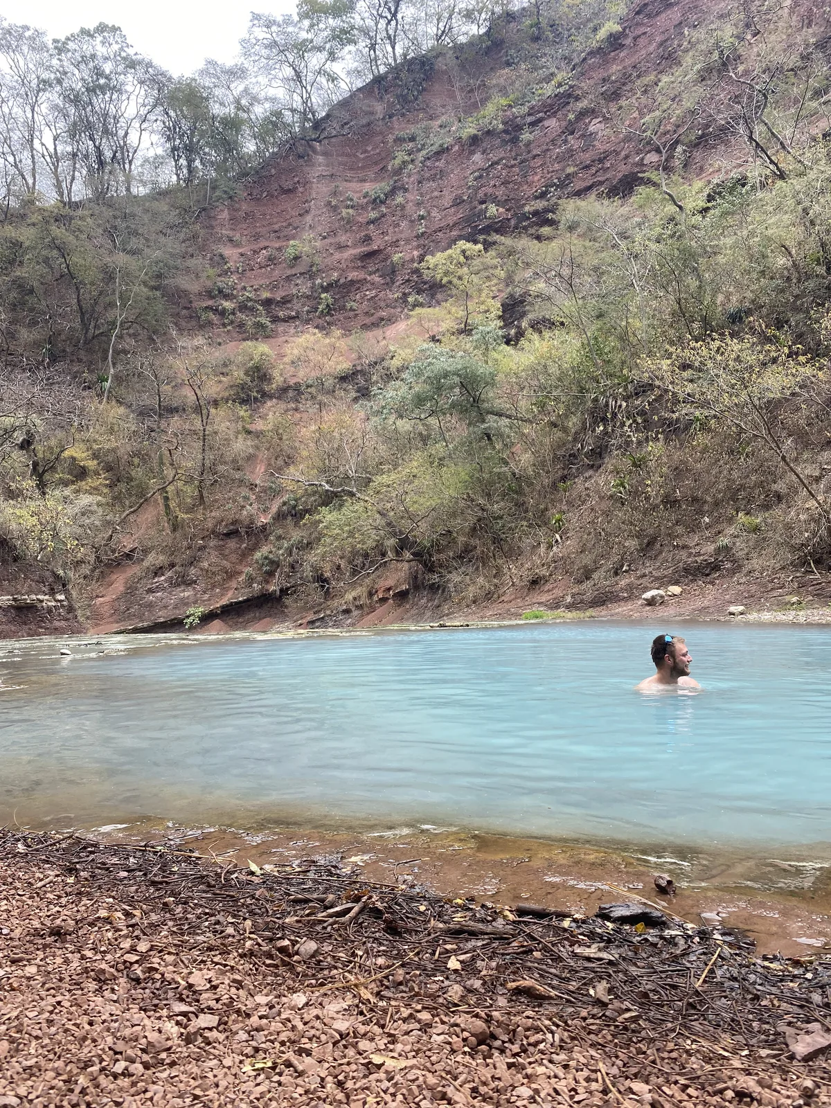 Hombre nadando en aguas azul celeste al pie de un acantilado de tierra rojiza, rodeado de vegetación tropical