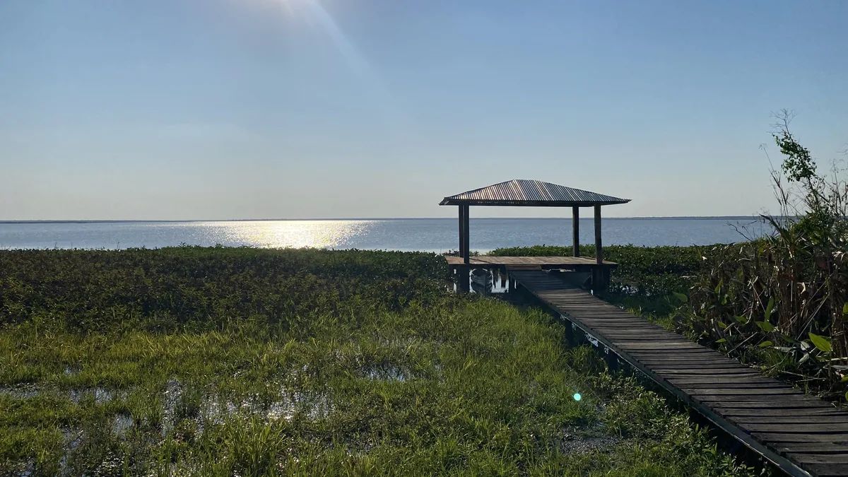 Reflejos del sol en un vasto lago o laguna con muelle de madera y gazebo, rodeado de exuberante vegetación acuática