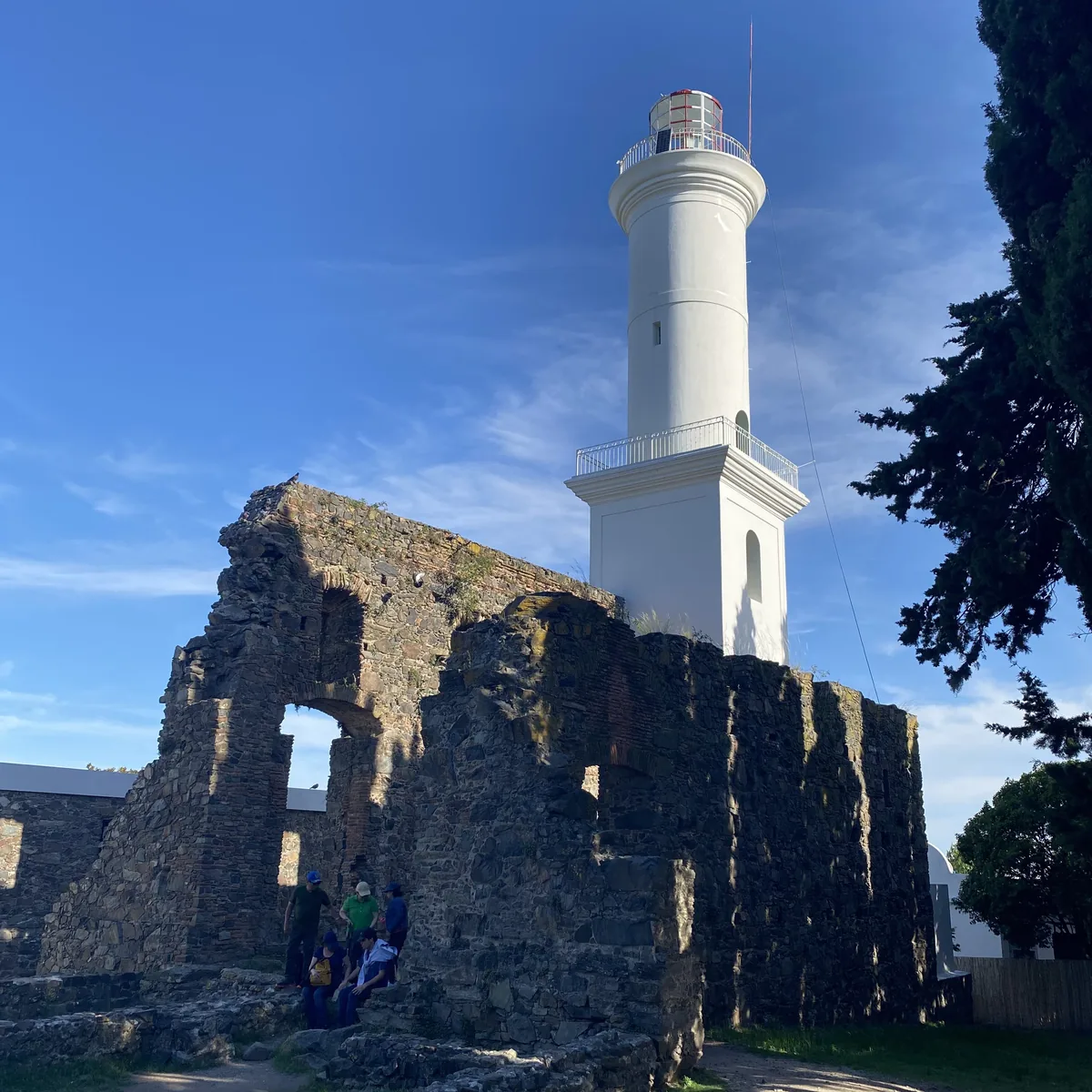 Faro blanco de Colonia del Sacramento sobre ruinas históricas de piedra, con viajeros explorando el sitio colonial bajo cielo
