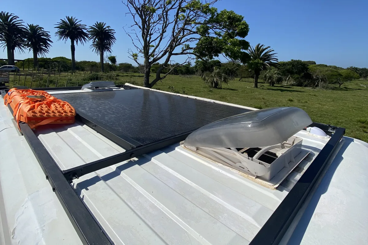 Techo de furgoneta overland con panel solar, ventilador de techo y tablas de desatasco naranjas, con palmeras y campo verde