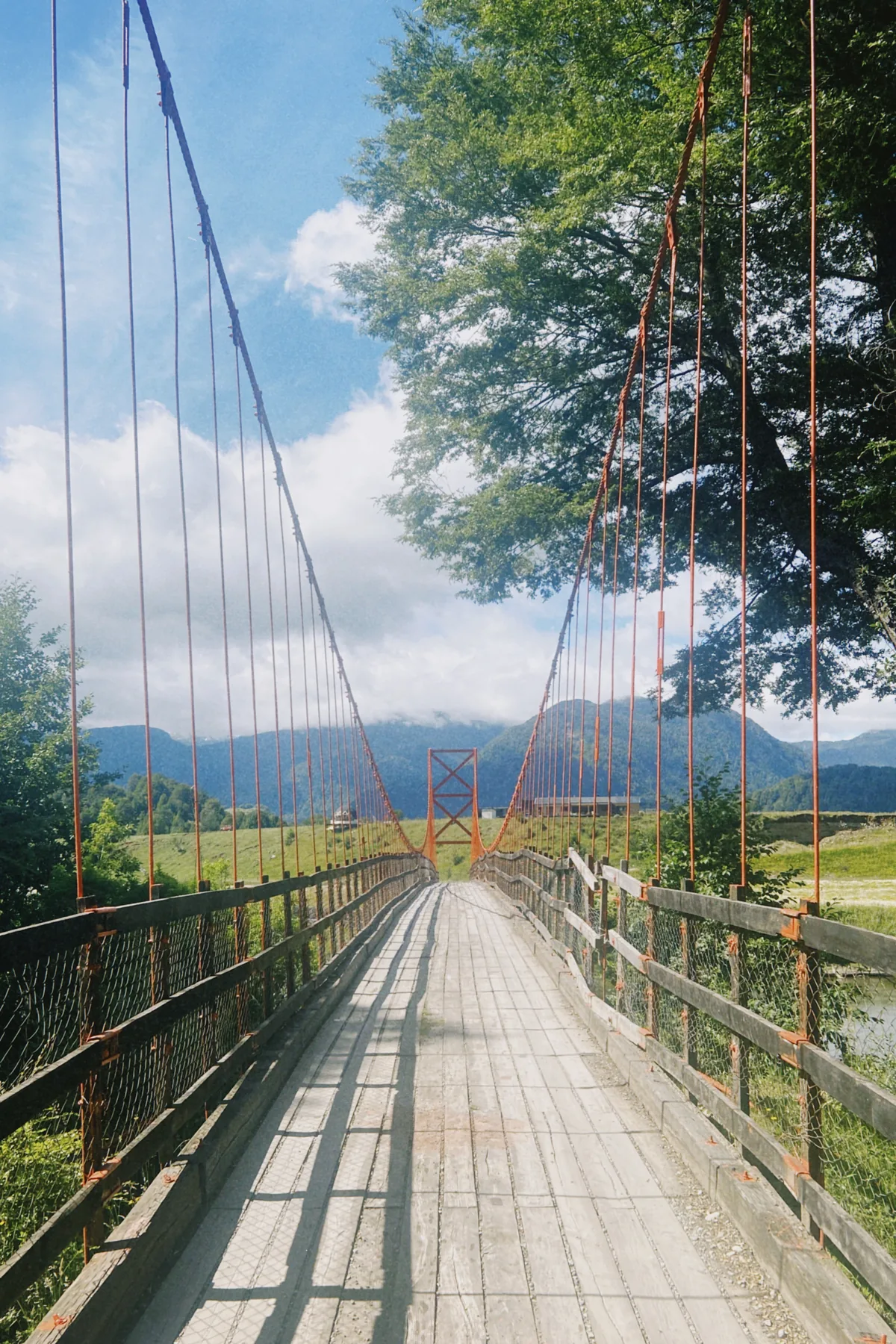 Cruce un pintoresco puente colgante de madera sobre un río, rodeado de frondosa vegetación y majestuosas montañas