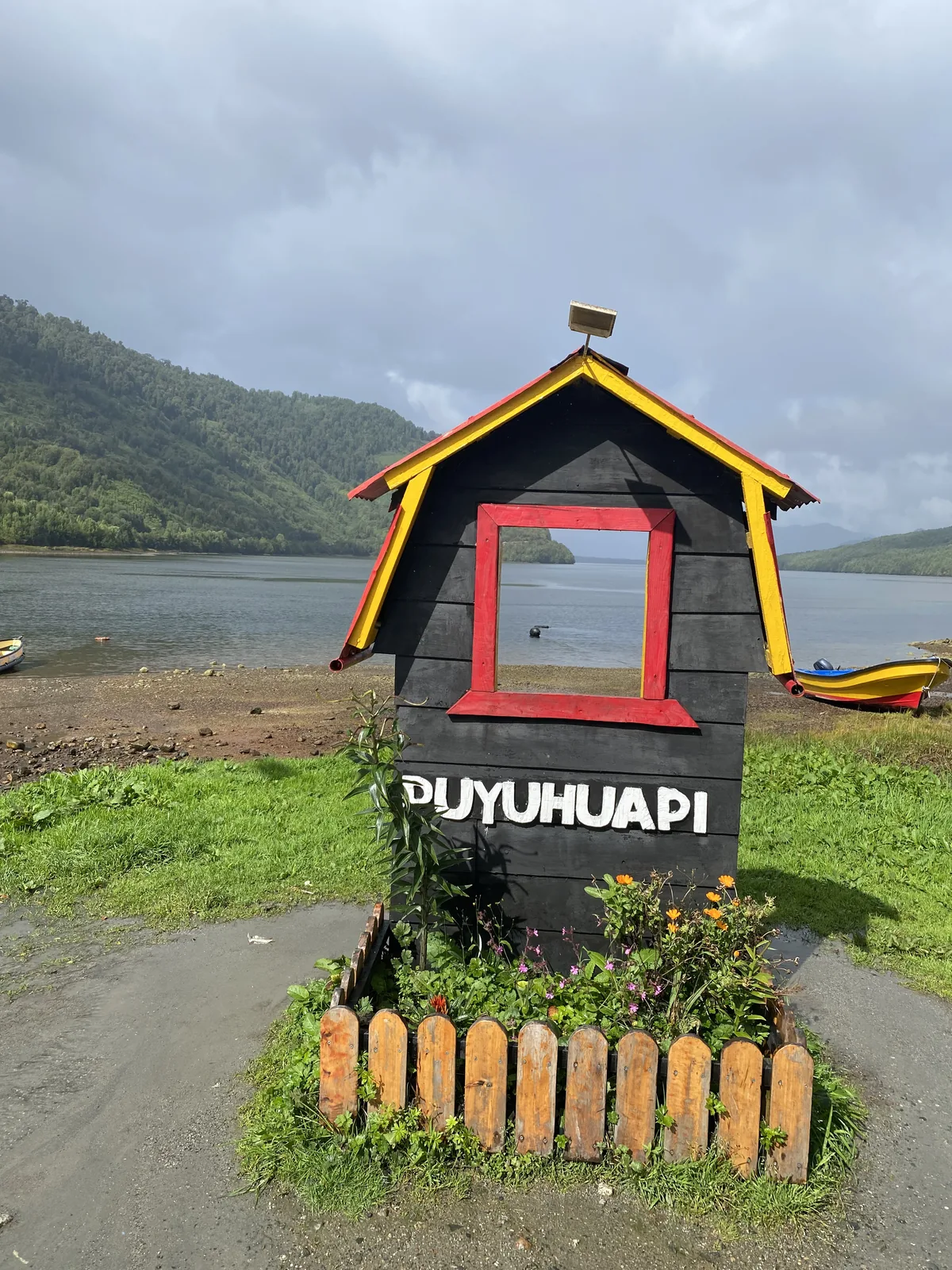 Cartel de bienvenida a Puyuhaipi con forma de casa frente a un fiordo, montañas verdes y cielo nublado en la Carretera Austra