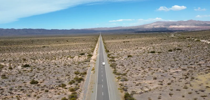 Furgoneta camper blanca en una ruta desértica y recta hacia el horizonte, rodeada de vastos paisajes áridos y sierras