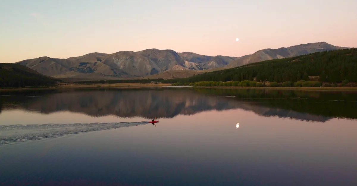 Kayaker en un lago alpino al atardecer, con montañas reflejadas en el agua tranquila y una luna brillante en el cielo