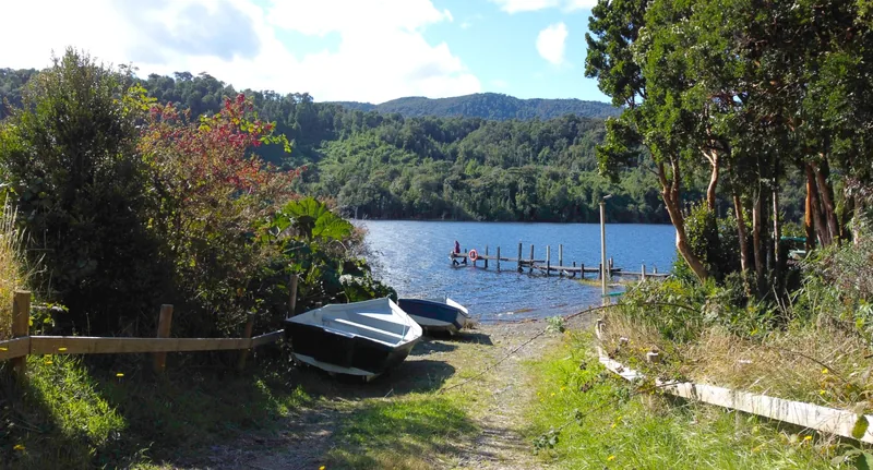 Persona en muelle de madera en un lago con dos botes en la orilla, rodeado de bosque andino y montañas bajo cielo azul