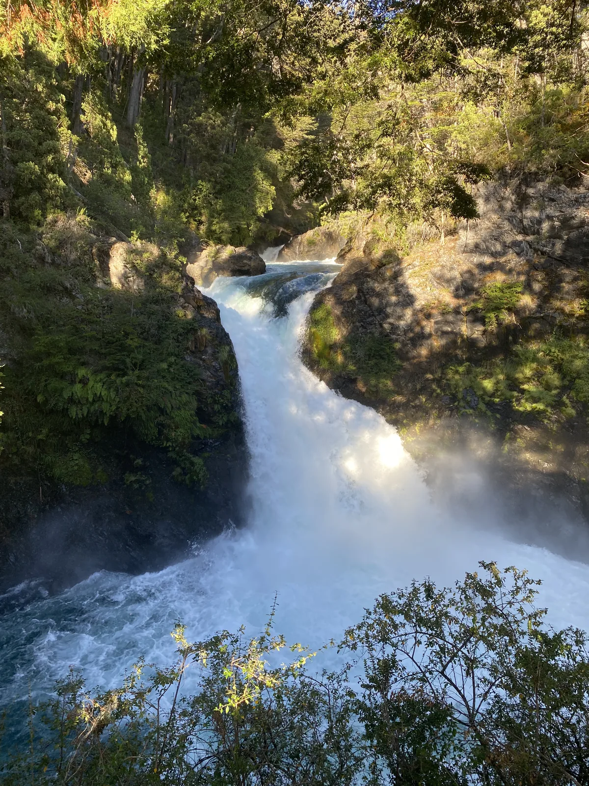 Poderosa cascada blanca cayendo entre paredes rocosas y bosque verde, con el sol iluminando el agua y la espuma