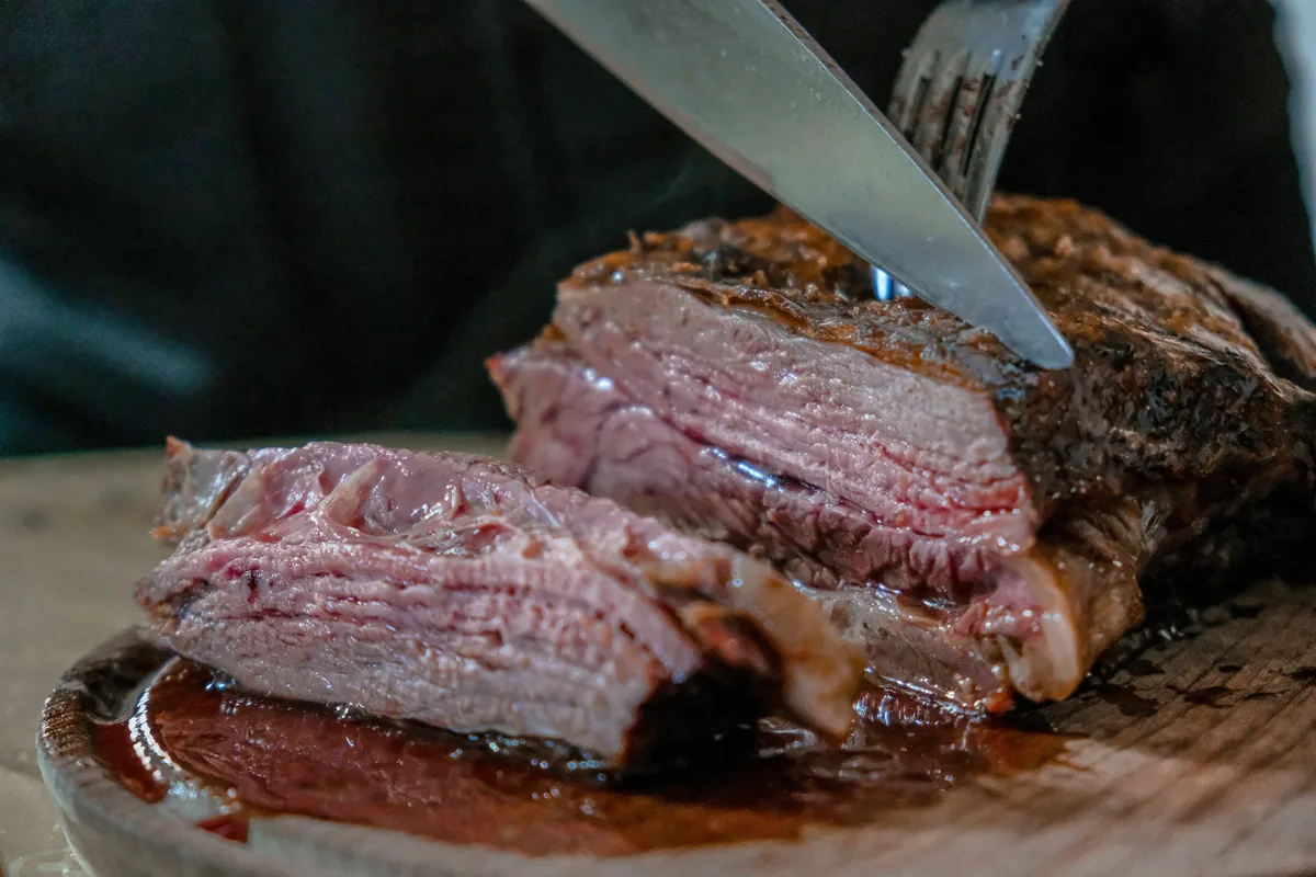 Jugosa carne asada al punto, con costra crujiente, siendo rebanada con cuchillo y tenedor en tabla de madera
