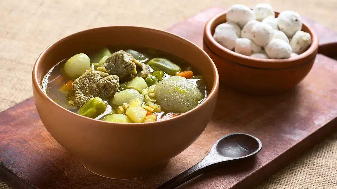 Caldo de carne con verduras andinas, papas y zanahorias en un cuenco rústico. Acompaña tazón de papitas andinas
