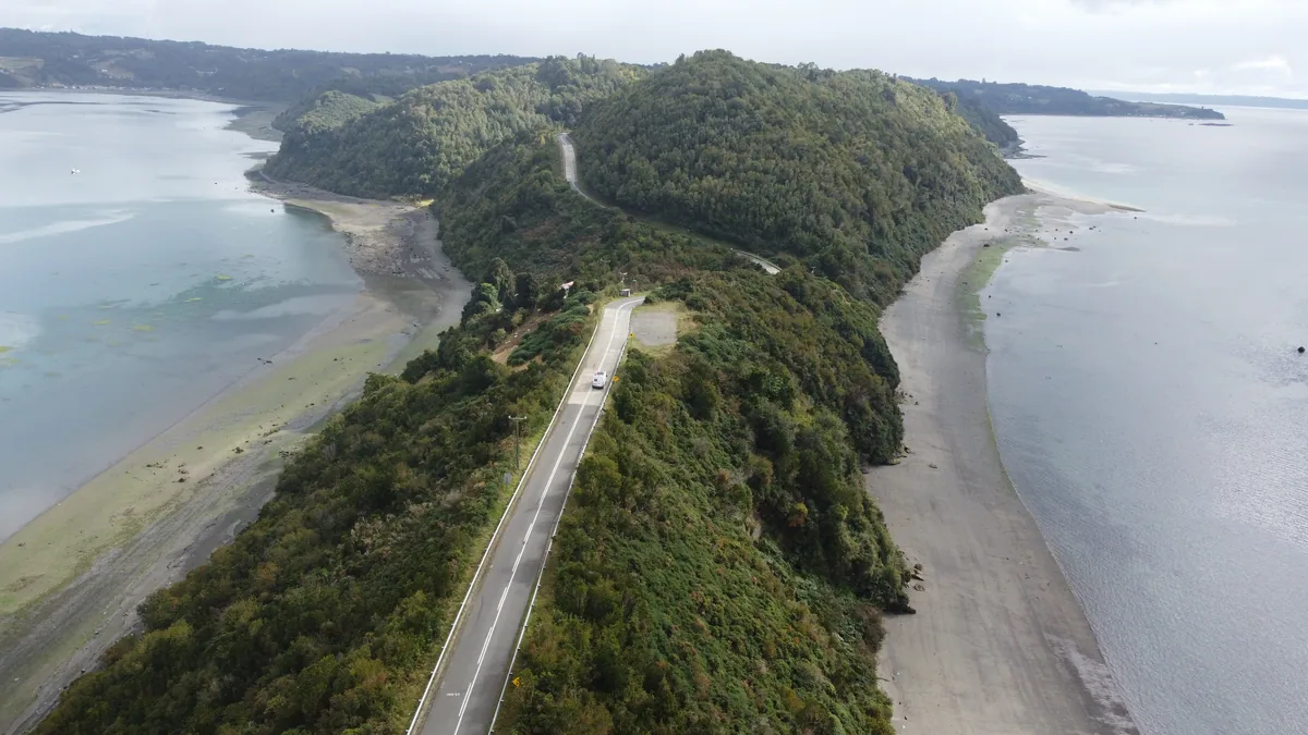 Furgoneta overland blanca recorre sinuosa carretera costera que cruza una península boscosa rodeada de bahías tranquilas