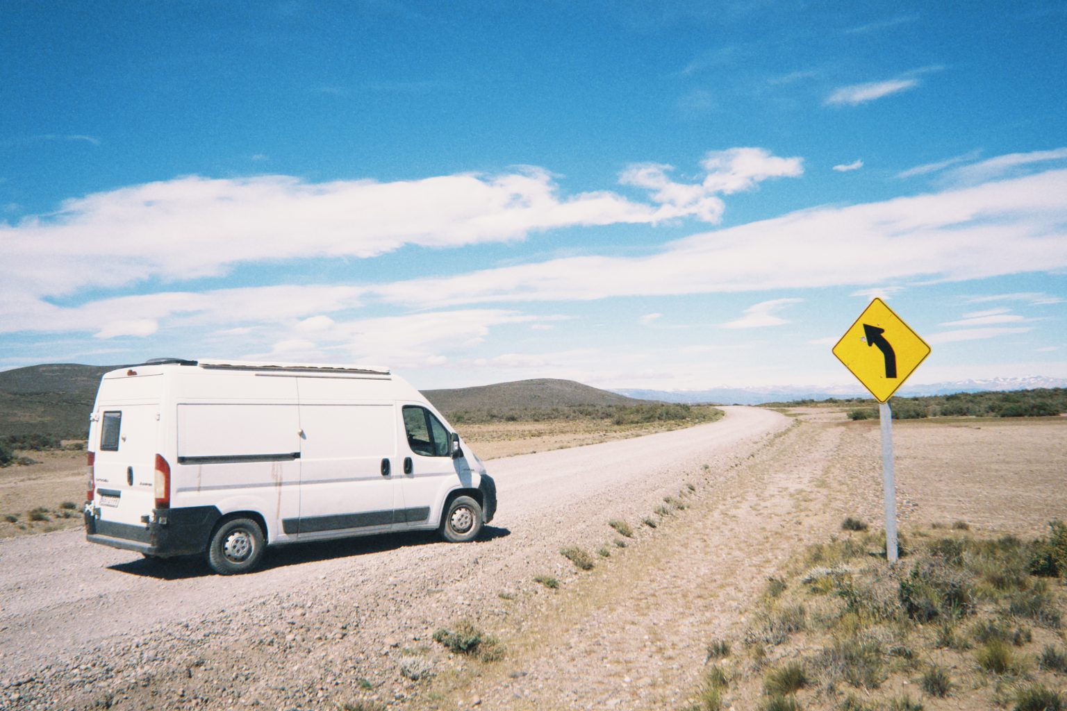 Van camper blanca sobre camino de ripio en la estepa patagónica, señal de curva a la izquierda y montañas lejanas