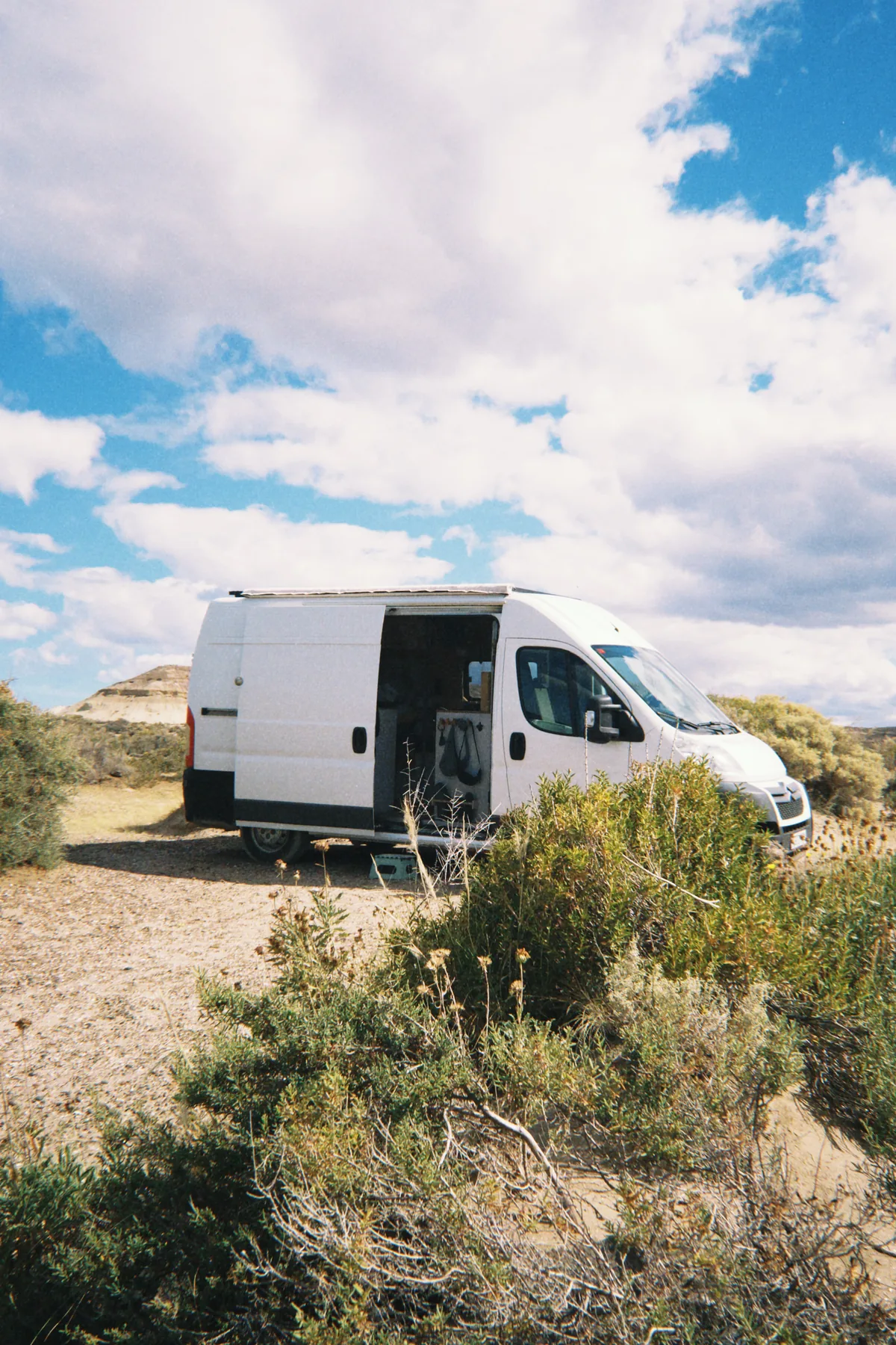 Furgoneta camper blanca con puerta abierta en un sitio de acampada en el desierto, entre arbustos y montañas