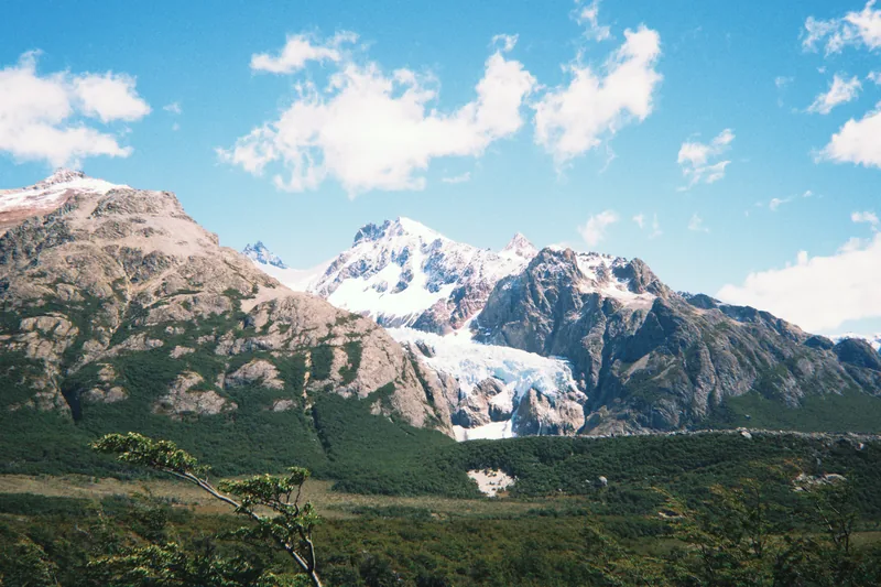 Montañas patagónicas con picos nevados y un glaciar azul, rodeadas de bosques verdes bajo el cielo azul con nubes