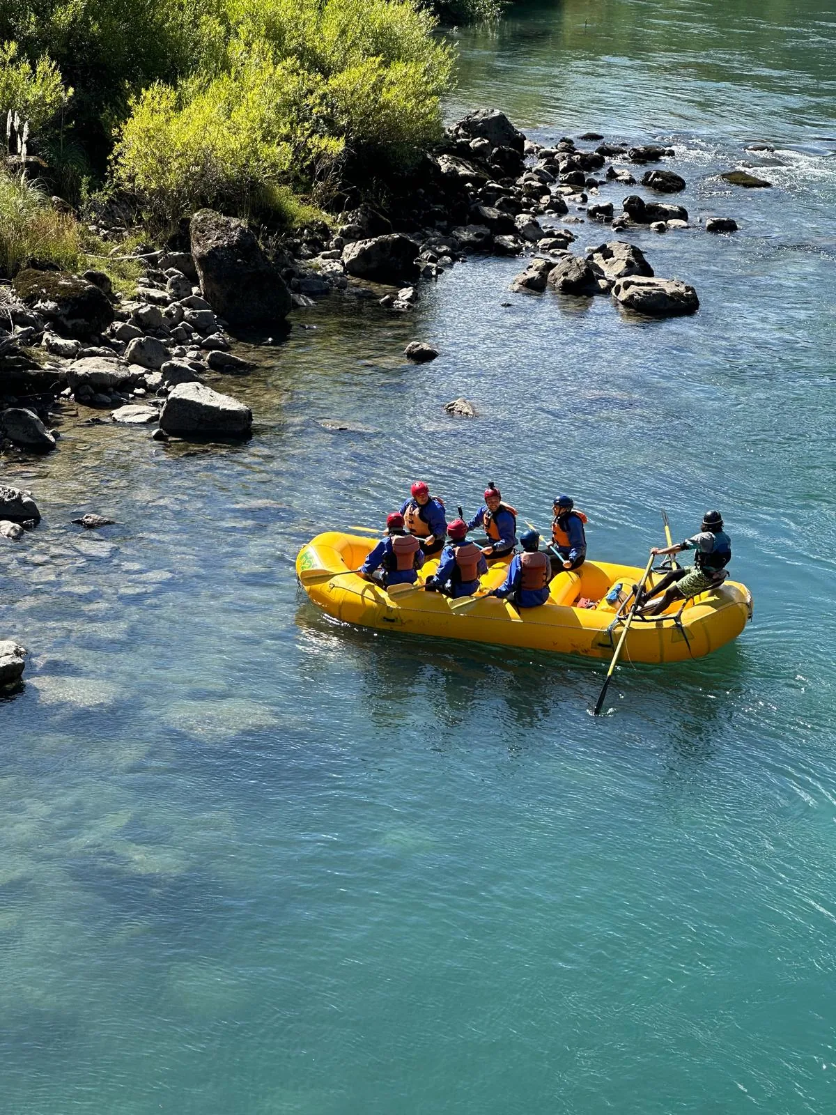 Balsa de rafting amarilla con 6 personas navegando río azul turquesa, junto a orilla rocosa con arbustos verdes