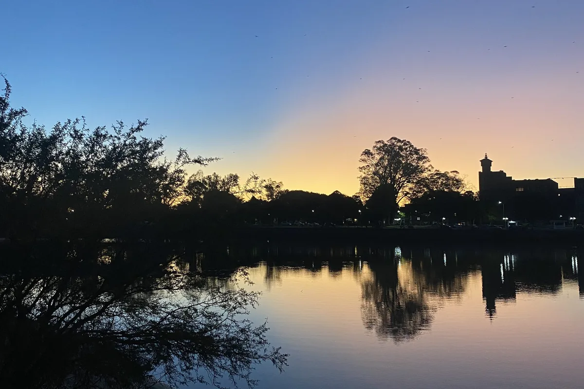 Silueta de árboles y edificios reflejándose en un lago al atardecer, con el cielo pintado de azules, naranjas y rosas