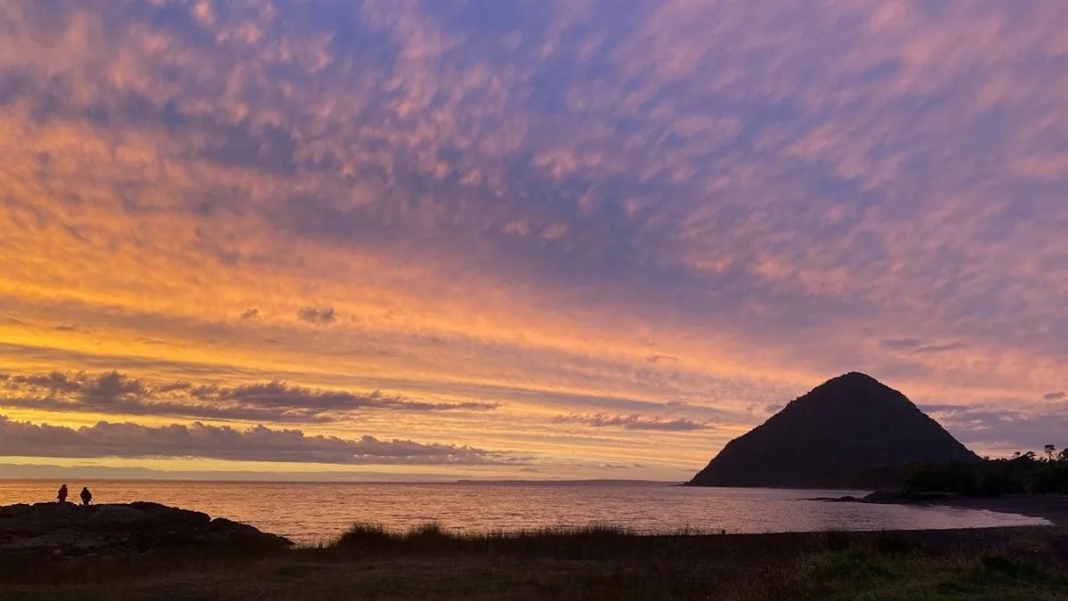 Espectacular atardecer con nubes rosas y naranjas sobre el mar, una montaña cónica y dos viajeros en la costa rocosa