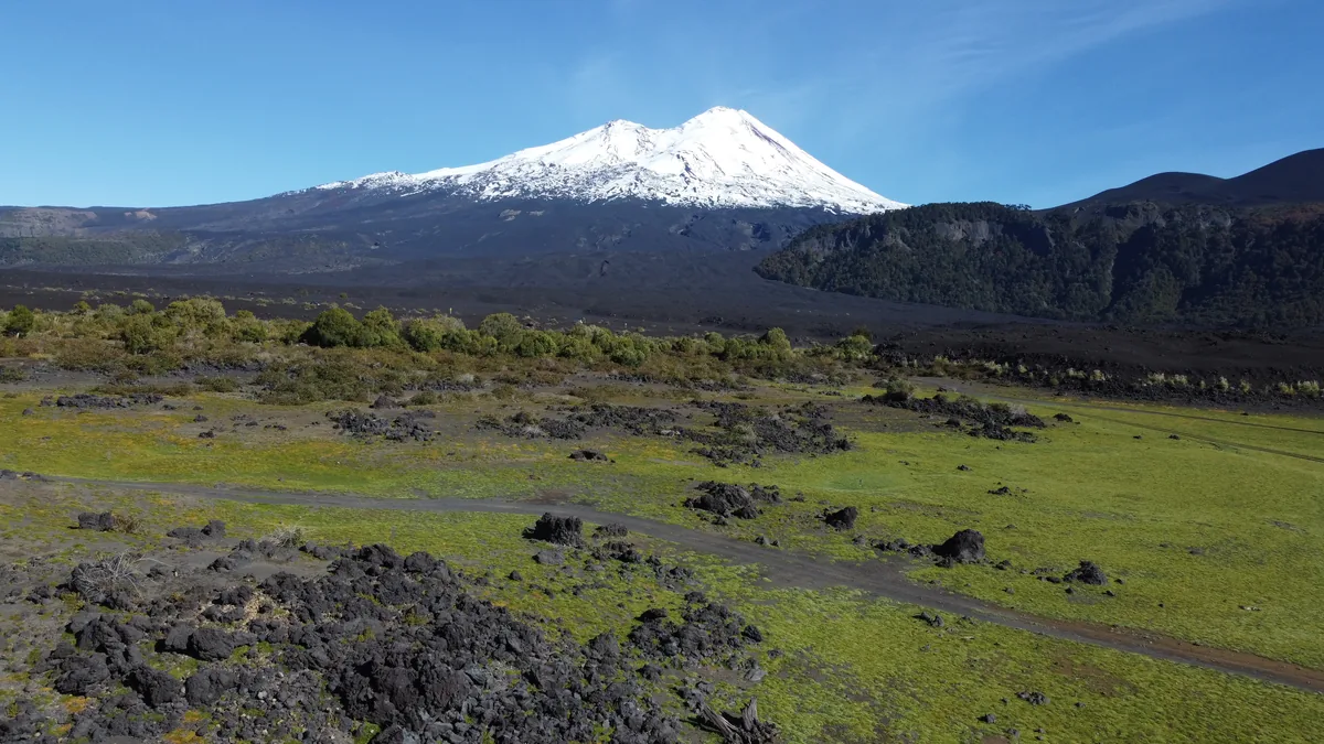 Volcán Llaima cubierto de nieve, flanqueado por campos de lava negra y senderos entre hierba verde y rocas volcánicas
