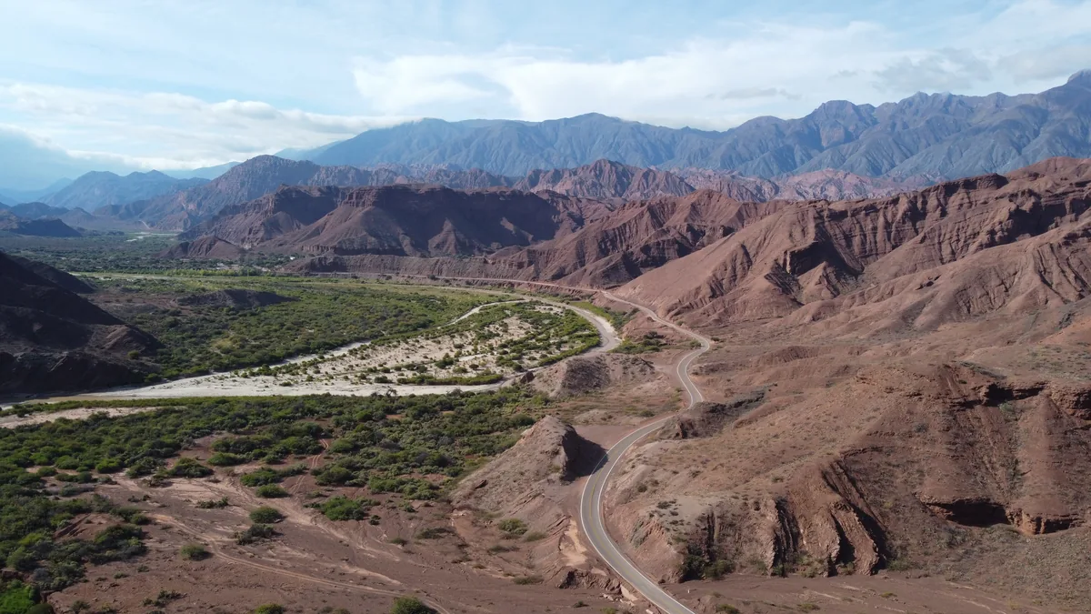 Carretera serpenteante entre imponentes montañas rojizas y un valle verde con río seco, bajo un cielo azul con nubes