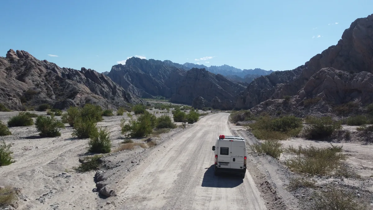 Furgoneta camper blanca con paneles solares avanzando por una ruta de tierra en un valle de montañas áridas y desérticas
