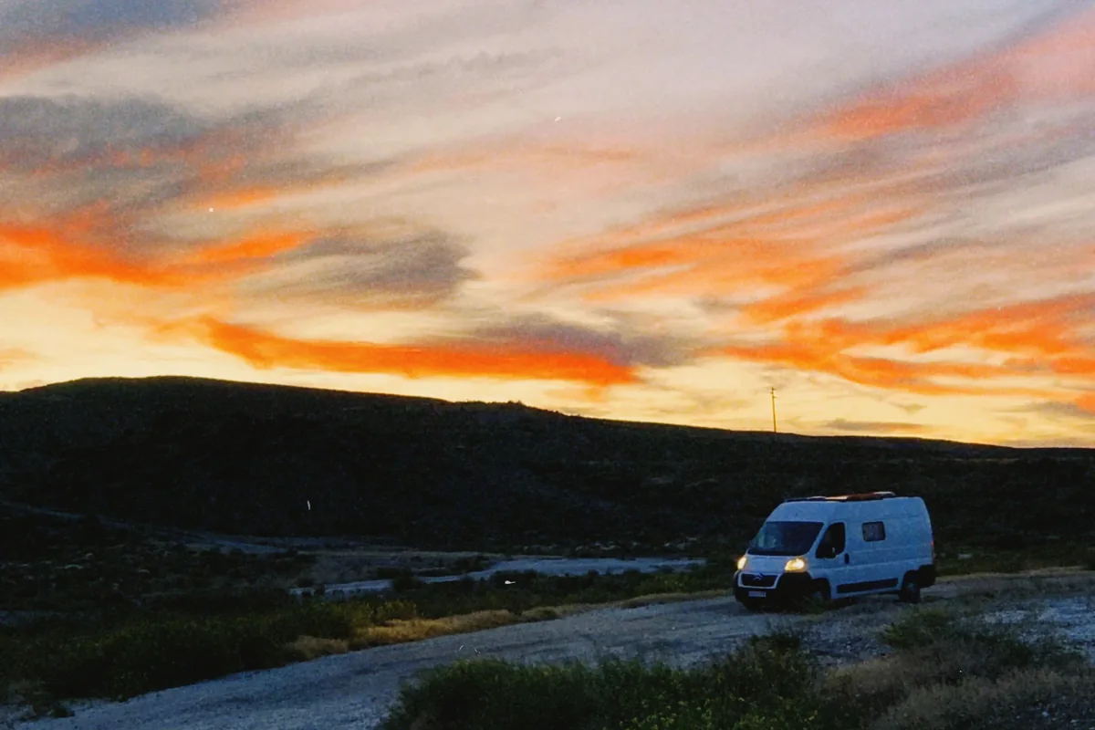 Van camperizada blanca con paneles solares en un camino de tierra al atardecer, con cielo naranja y montañas oscuras