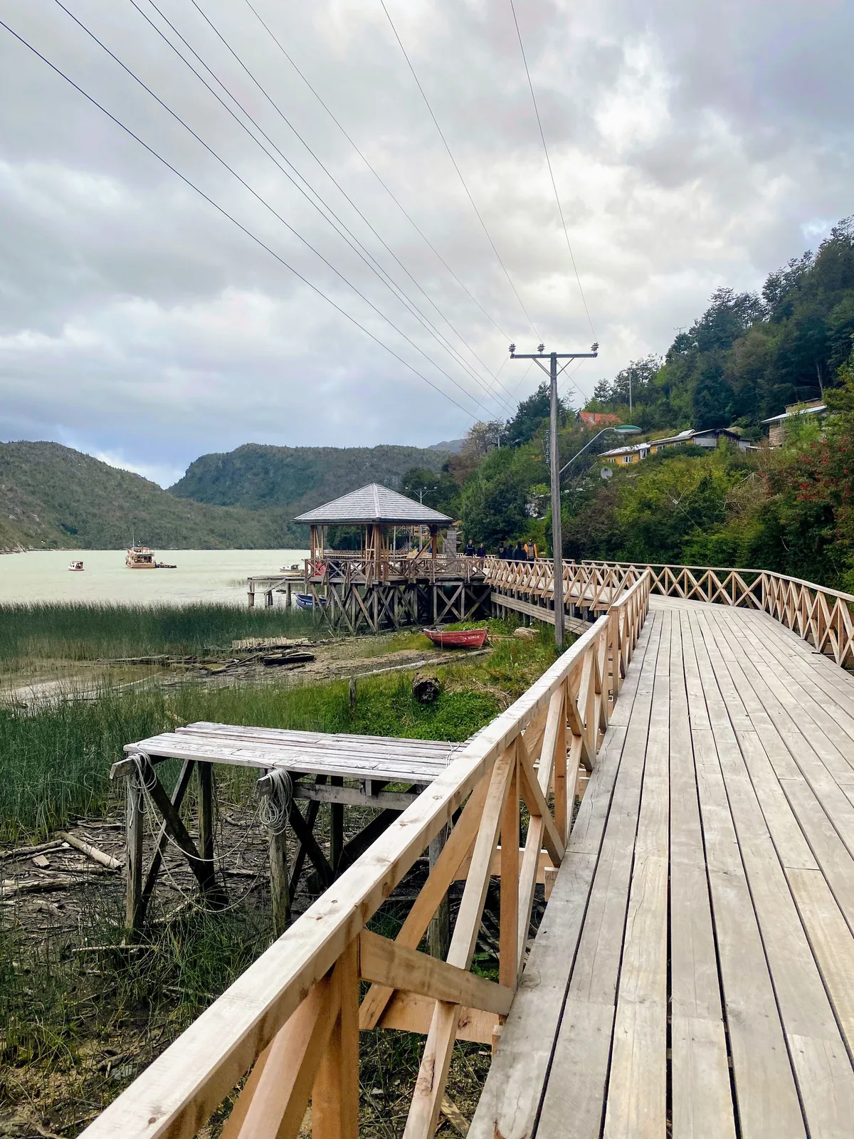 Pasarela de madera que bordea una bahía de aguas turquesas con un gazebo sobre el agua, entre montañas verdes
